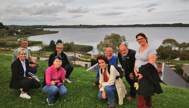 Projekta "Shared Spaces" dalībnieki – Ludzas un Buržas pašvaldību pārstāvji – smaidot iepozē zaļā pakalnā, fona redzama Ludzas ezera ainava un jaunizbūvētā promenāde.