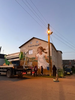 Murāļa "Es atgriežos Latgalē" tapšanas process pievakarē: pie ēkas sienas novietots pacēlājs un mākslinieki strādā pie sienas gleznojuma izstrādes.
