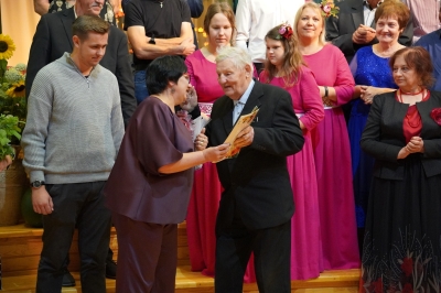 Pušmucovas Tautas nama vadītāja Inta Domarka pasniedz pateicības rakstu vecākajam saieta muzikantam Leonam Bērziņam 2024. gada tautas muzikantu saietā Pušmucovā, fona dalībniekiem smaidot. Fotogrāfiju autore: Jana Slokenberga.