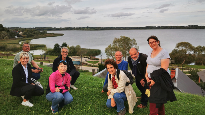 Projekta "Shared Spaces" dalībnieki – Ludzas un Buržas pašvaldību pārstāvji – smaidot iepozē zaļā pakalnā, fona redzama Ludzas ezera ainava un jaunizbūvētā promenāde.