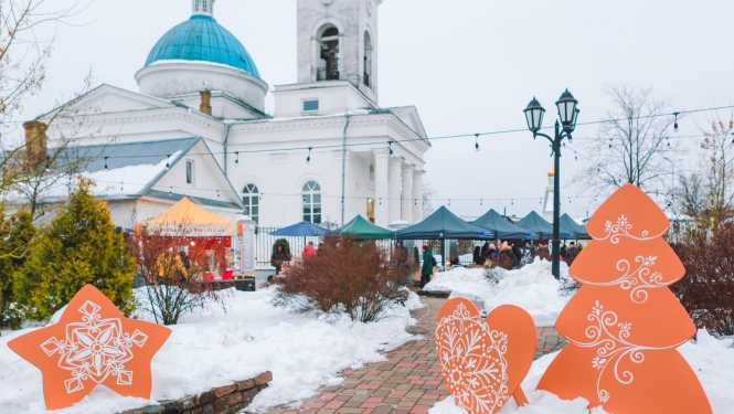 Ziemassvētku tirdziņš ar tirdzniecības teltīm sniegotā Vecpilsētas laukumā Ludzā, fonā Dievmātes Patvēruma pareizticīgo baznīca, priekšplānā oranži svētku dekori. Foto: Ralfs Punans.