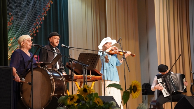 Zvirgzdenes lauku kapela uzstājas tautas muzikantu saietā Pušmucovā 2025. gadā. Uz skatuves redzami muzikanti ar bungām, vijoli, akordeonu un citiem instrumentiem, priekšplānā saulespuķu dekori. Fotogrāfiju autore: Jana Slokenberga.