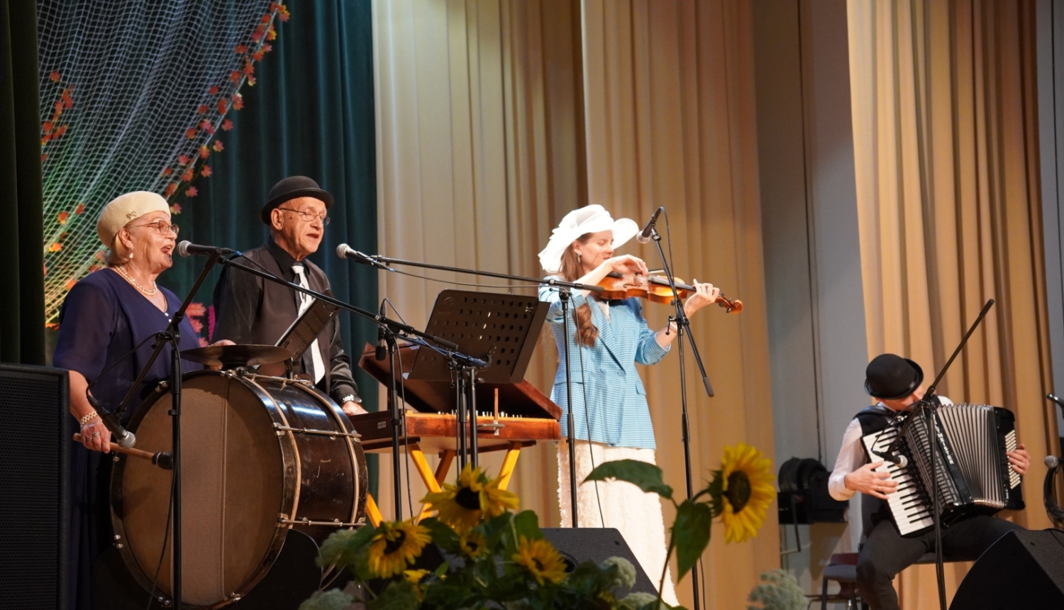 Zvirgzdenes lauku kapela uzstājas tautas muzikantu saietā Pušmucovā 2025. gadā. Uz skatuves redzami muzikanti ar bungām, vijoli, akordeonu un citiem instrumentiem, priekšplānā saulespuķu dekori. Fotogrāfiju autore: Jana Slokenberga.